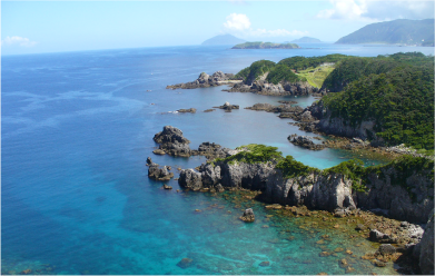 The emerald green sea stretches out along the coast of Shikine-jima in the Izu Islands. The rocky coastline is accompanied by lush green hills, and islands can be seen in the distance.
