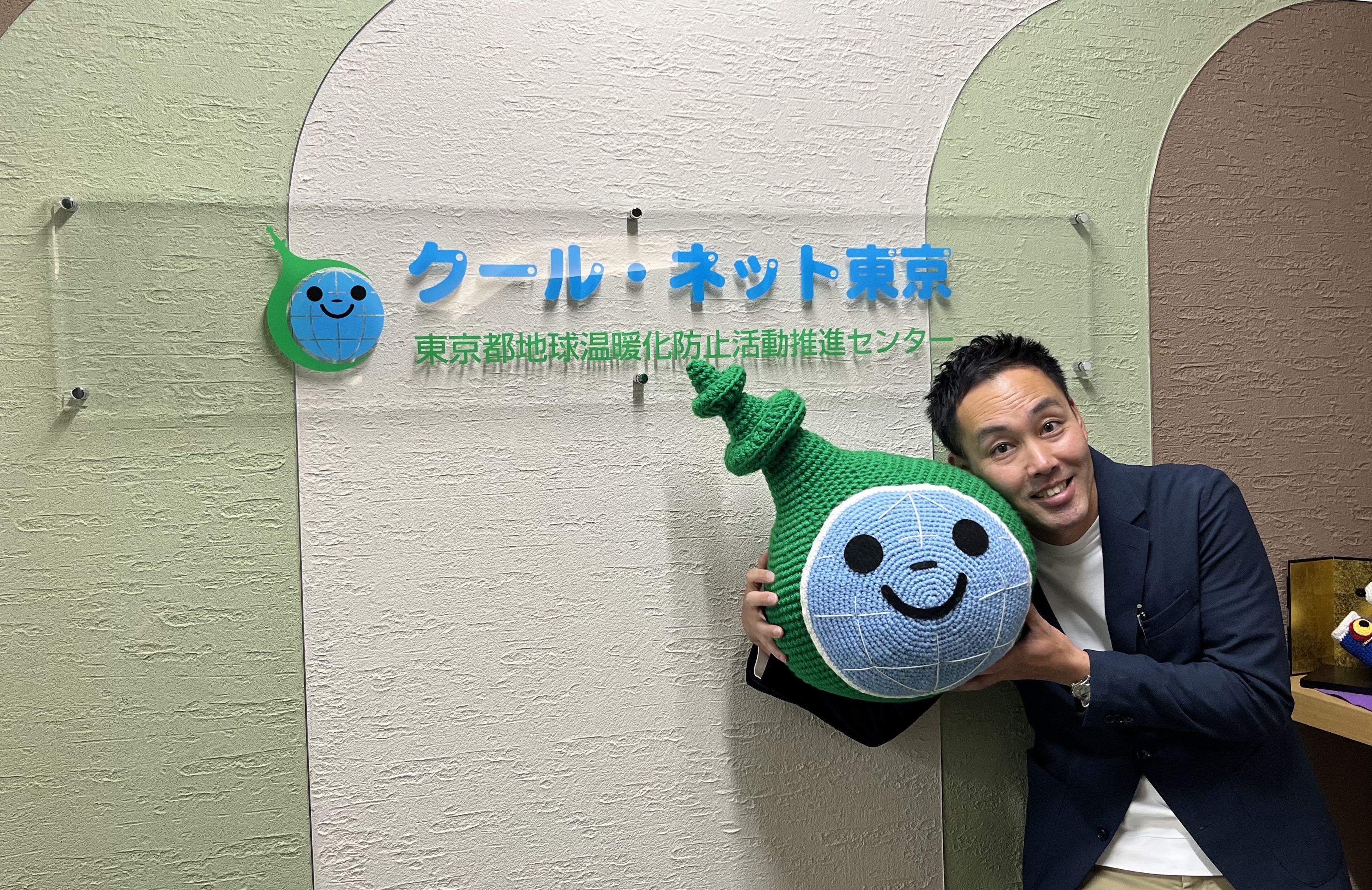 クール・ネット東京の受付で米田さんが人形を持って笑っている写真